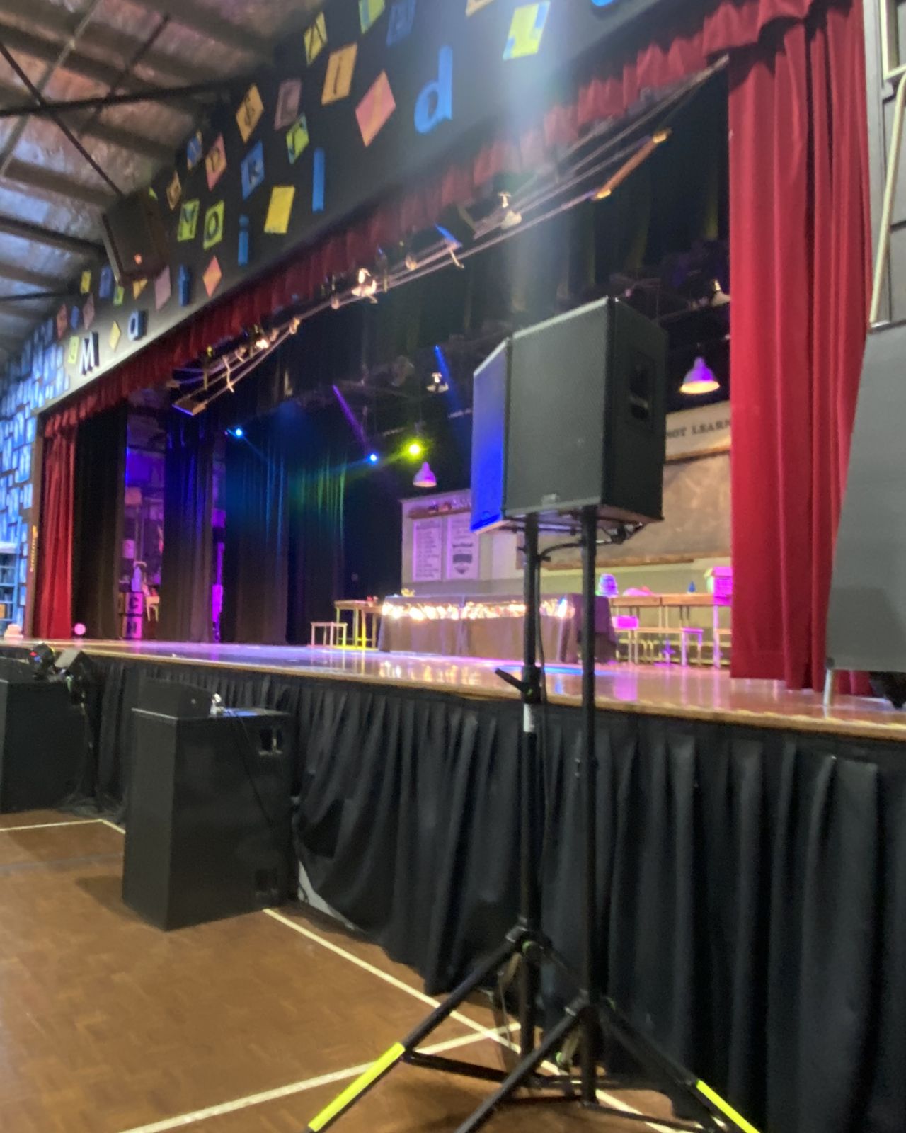 School hall set for a production, PA speakers on stands beside the stage